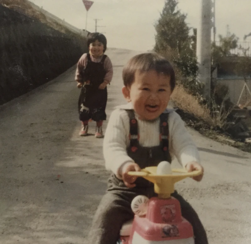 在未鋪柏油的坡道上騎著三輪車笑著的幼年時期的我,後面站著姊姊