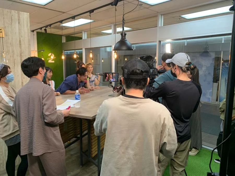 辦公室裡的拍攝現場。工作人員架著攝影機與收音設備,拍攝坐在長桌旁受訪的人